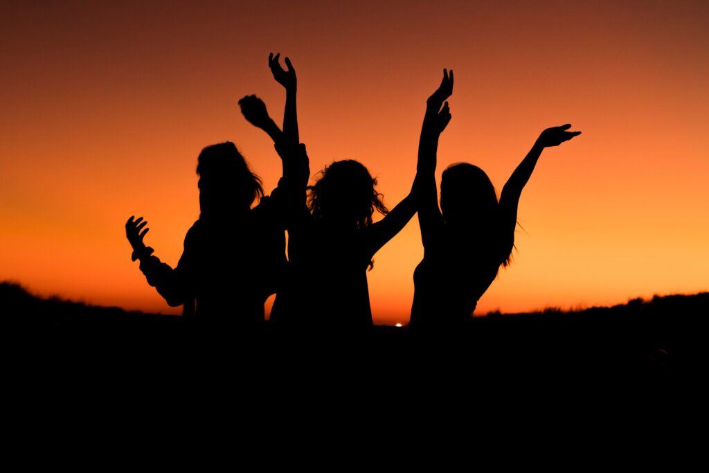 Three women are standing in the sunset with their arms up.